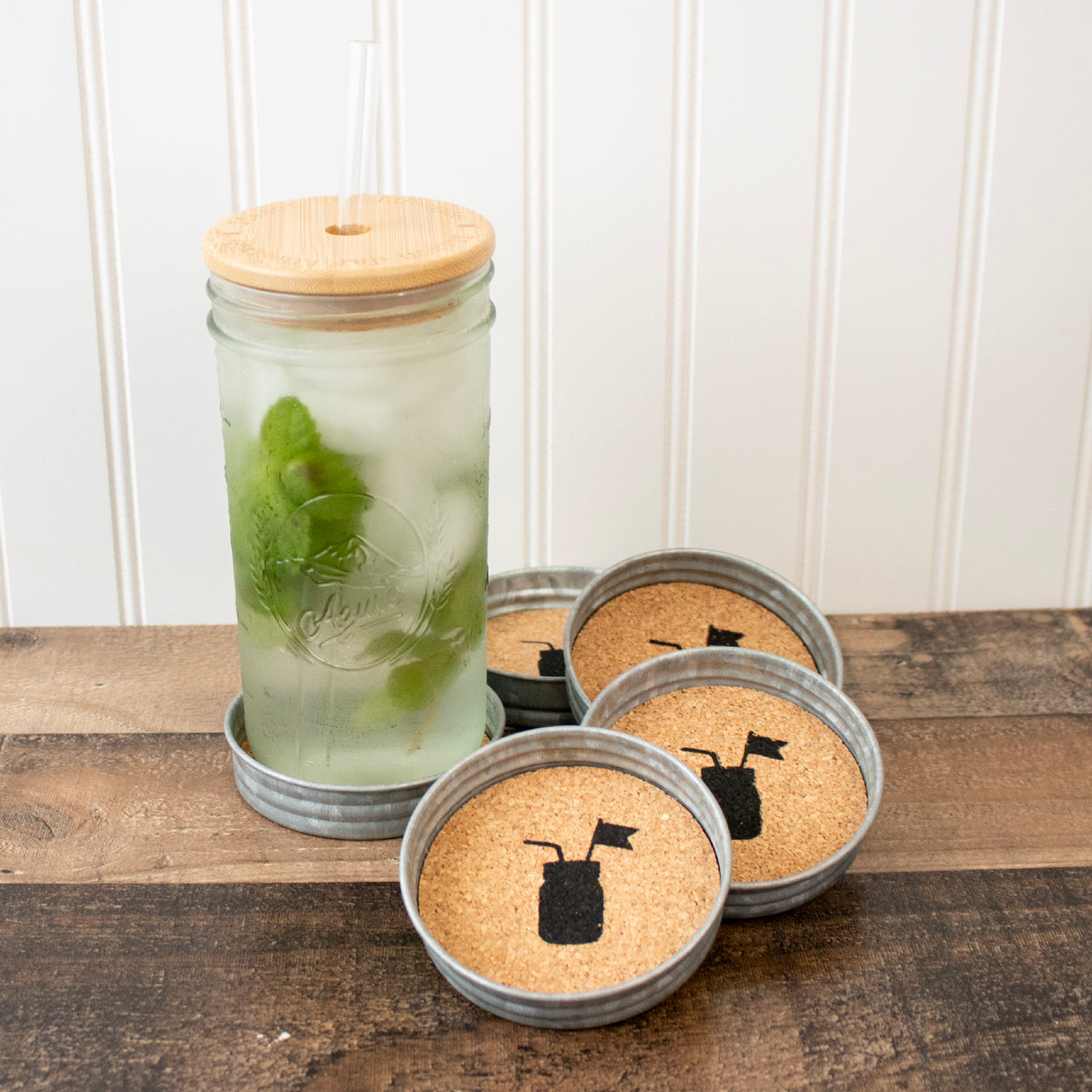 Four logo galvanized Mason jar lid-style coasters with cork inserts, shown with a Mason jar containing a drink and mint leaves.