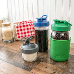 Charcoal Gray, Deep Blue, and Leaf Green Pour & Store shaker lids with carry loops on filled Mason jars on a kitchen counter.
