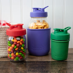 Cherry Red, Ultra Violet, and Leaf Green Pour & Store shaker lids with carry loops, shown on filled Mason jars with silicone sleeves.
